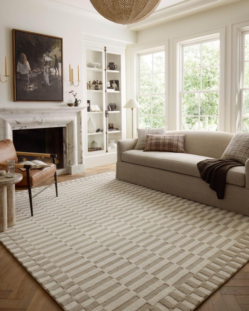 Cozy living room with a light beige sofa, a warm leather armchair, and a patterned area rug. A fireplace and built-in shelving are visible, with large windows allowing natural light to enter the space.