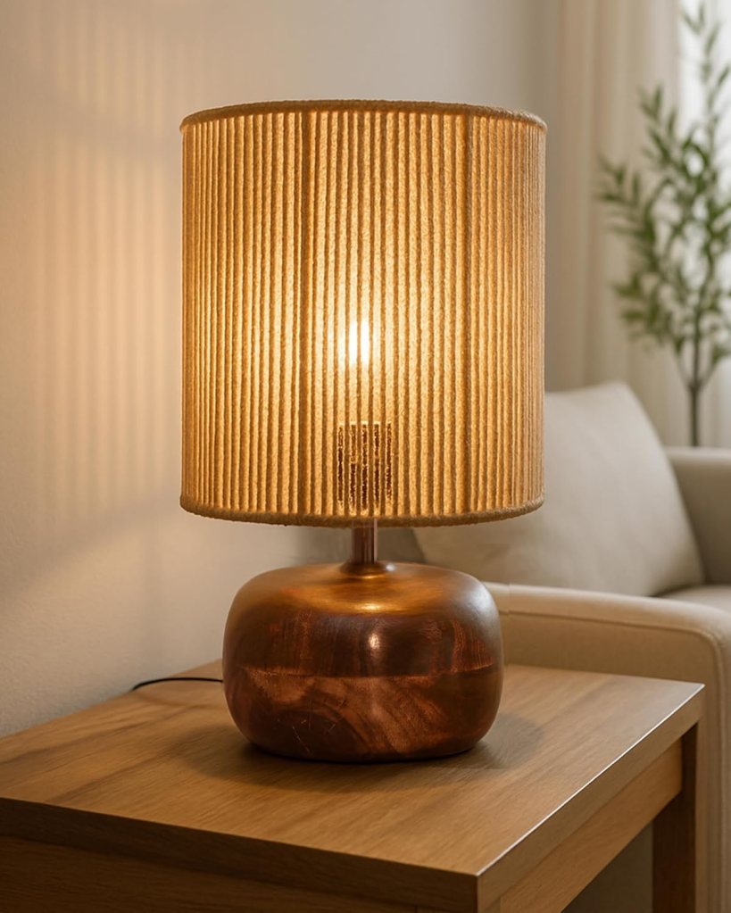 Table lamp with a beige, pleated shade and a round, copper base, emitting warm light in a cozy living room setting.